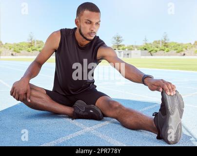 Corridore maschile che fa esercizi di stretching mentre è seduto su una pista sportiva. Giovane atleta maschile determinato seduto da solo e riscaldando i muscoli prima di andare a correre. Uomo che si prepara per una gara Foto Stock