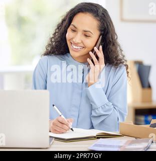 Il multitasking verso il successo. Scatto di una giovane donna d'affari che scrive le note mentre parla su un cellulare e usando un laptop in un ufficio. Foto Stock