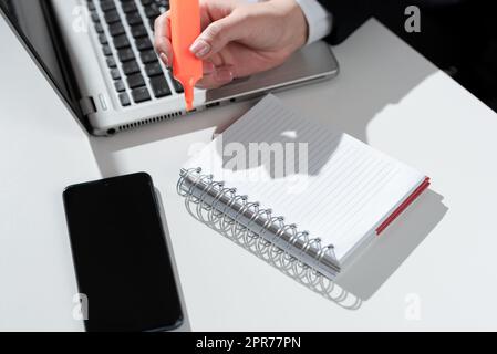 Donna d'affari che scrive nel notebook con il contrassegno sulla scrivania con il telefono cellulare e il computer portatile. Donna in tuta che tiene la penna colorata sopra il blocco note sul tavolo con cellulare e computer. Foto Stock