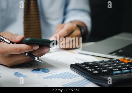 Uomo d'affari che tiene il telefono cellulare e la penna con grafici e grafici documenti aziendali sulla scrivania. Calcolatrice e computer per gli investimenti e l'analisi economica. Concetto di business finanziario e fiscale. Foto Stock