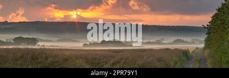 Vista panoramica della nebbia su campo remoto con spazio di copia al tramonto. Nebbia che copre una vasta distesa di prati di campagna in Germania all'alba. Fumo proveniente da incendi boschivi o incendi boschivi che attraversano la riserva naturale Foto Stock