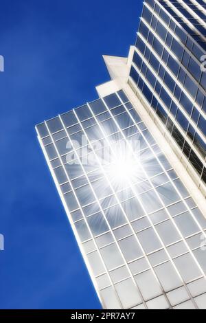 Finestre geometriche in vetro su un grattacielo con il sole che riflette un riflesso della lente su uno sfondo blu del cielo dal basso. Dettagli architettonici esterni di un alto e moderno edificio in città Foto Stock