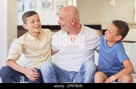 Uomo caucasico e i suoi due figli seduti sul divano a casa. Felice famiglia di tre persone in soggiorno. Due fratelli seduti con il loro padre. Sorridere, legare e passare del tempo insieme Foto Stock