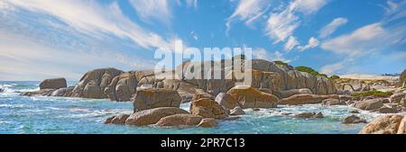 Vista panoramica sul mare di Camps Bay, città del Capo, Sud Africa con massi, rocce e cielo blu con spazio fotocopie. Le onde oceaniche si gettano su una spietata spiaggia rocciosa. Viaggi e turismo all'estero e all'estero Foto Stock