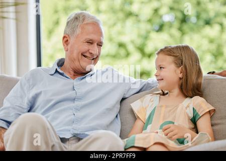 Bell'anziano e sua nipote seduti su un divano nel soggiorno di casa. Bambina in visita al suo nonno. Legare con un nipote e' un momento molto speciale nella vita Foto Stock