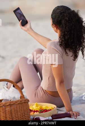 Catturare alcuni dei miei momenti preferiti sulla spiaggia: Una donna irriconoscibile seduta da sola sulla spiaggia e usando il cellulare per prendere selfie. Foto Stock