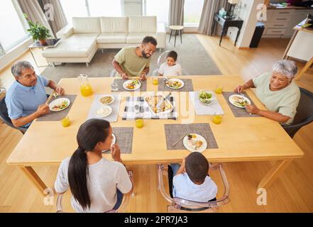 Vista dall'alto di una famiglia da corsa mista seduta a un tavolo per pranzare nel salotto di casa. Nonni ispanici che hanno un pasto con i loro figli e gr Foto Stock