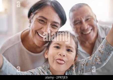 Ritratto di sorridenti nonni e nipoti di razza mista che scattano un selfie nella sala di casa. Uomo e donna anziano ispanico che scattano foto e legano con la loro piccola nipote carina a casa Foto Stock