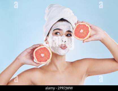 Bellissima giovane donna mista che indossa una maschera facciale e un asciugamano mentre posa con un pompelmo. Bella donna pulita e fresca fuori dalla doccia e applicando il suo regime giornaliero di cura della pelle sul viso Foto Stock