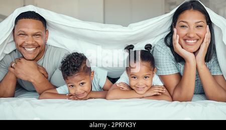 Ritratto di una famiglia felice con due bambini sdraiati sotto il piumone che sorridono e guardano la macchina fotografica. Bambina e ragazzo sdraiati a letto con i loro genitori che legano e si divertono Foto Stock