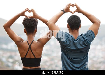 La coppia afro-americana con vista posteriore crea una forma del cuore con le mani mentre si esercita all'aperto. Giovane atletico uomo e donna che promuove la salute e la forma fisica all'aperto. Amano allenarsi insieme Foto Stock