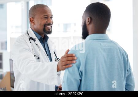 Un medico maturo che fornisce supporto al paziente. Un medico afroamericano che tocca un paziente sul braccio. Paziente in un consulto con il suo medico. Un medico curante che offre comfort e supporto al paziente. Foto Stock