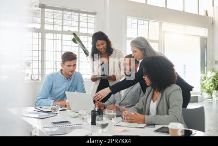 Felice e diversificato gruppo di uomini d'affari che si riuniscono mentre si trovano a un tavolo al lavoro. Professionisti che parlano e pianificano mentre utilizzano un notebook in ufficio. Colleghi di sesso maschile e femminile che discutono di una strategia aziendale Foto Stock