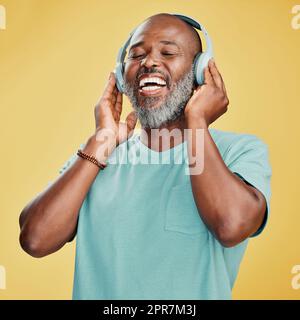 Felice uomo afroamericano maturo, in piedi da solo su uno sfondo giallo in uno studio di registrazione e indossa cuffie per ascoltare musica. Ritratto sorridente di un uomo nero anziano con barba grigia che ascolta la musica Foto Stock