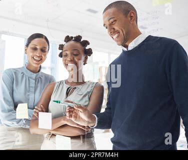 Tre persone d'affari felici e diversificate che scrivono idee e collaborano in un ufficio al lavoro. Allegro uomo d'affari ispanico che fa una nota su una finestra di vetro con colleghi donne Foto Stock