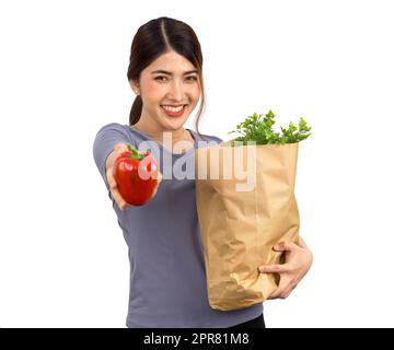 Giovane donna asiatica in abbigliamento casual con un sorriso, offre il peperone rosso sul davanti mentre tiene il sacchetto di carta pieno di verdure. Ritratto su sfondo bianco con luce da studio. Isolato Foto Stock