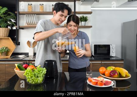 Un giovane vestito casual versa frutta mista e frullato vegetariano dalla macchina per frullare nel bicchiere della sua fidanzata. Gli amanti asiatici trascorrono la mattina insieme in cucina. Foto Stock