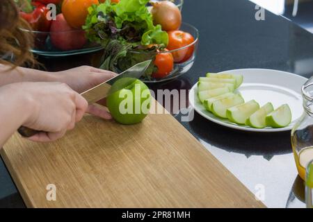 Chiudi la mano tenendo il coltello che taglia la mela verde su un tagliere di legno. La frutta a fette viene posta su un piatto. Una ciotola di vetro con una varietà di frutta e verdura è posizionata sul bancone della cucina. Foto Stock