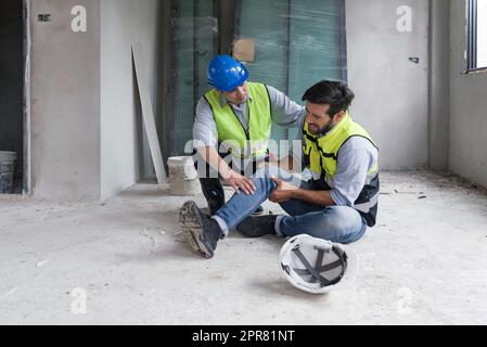 Il giovane uomo caucasico in giubbotto di sicurezza gli afferrò il ginocchio e urla nel dolore. A causa di lesioni in cantiere. Il suo amico asiatico viene e lo aiuta a stare in piedi. Foto Stock