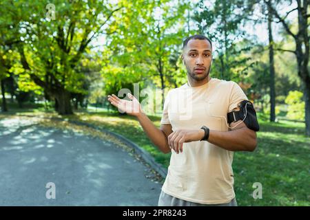 Maratona sportiva, gara. Un giovane afroamericano che si trova in piedi nel parco e guarda con parole preoccupate l'orologio intelligente, il braccialetto fitness, frustrato con il tempo, le mani agitate alla macchina fotografica. Foto Stock