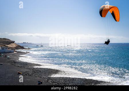 Mentre il sole splende sulla spiaggia di Playa de la Enramada, una popolare zona di atterraggio del parapendio a Tenerife, un parapendio irriconoscibile sta atterrando. Foto Stock