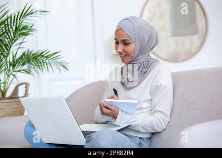 Apprendimento a distanza. Giovane donna latino-americana in hijab studente di studio on-line. Si siede a casa sul divano con un computer portatile in grembo e scrive una lezione, una lezione, un corso in un notebook. Foto Stock