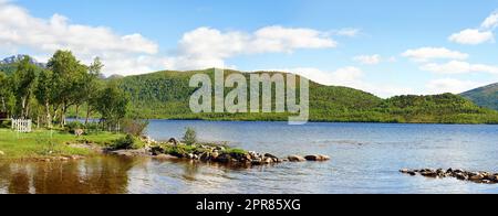 Paesaggio di un lago con alberi, verdi colline erbose. Un suggestivo pozzo d'acqua circondato da un terreno boschivo vuoto. Costa del lago di montagna con cielo nuvoloso blu nel Nordland vicino a Bodo, Norvegia Foto Stock