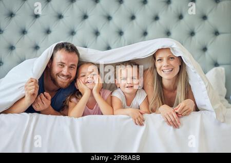 Una famiglia felice a letto sotto una coperta a casa. Ritratto di giovani genitori sorridenti che si divertono con i bambini in camera da letto, coperto da un piumone. Bambine carine che giocano con la madre e il padre Foto Stock