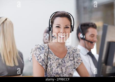 Come posso aiutarla. Un ottimo rappresentante del servizio clienti sorride alla fotocamera con i colleghi che lavorano in background. Foto Stock