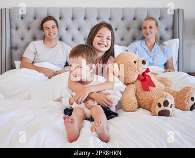 Adorabile bambina che abbraccia il fratello minore con il suo orsacchiotto accanto a lei mentre è seduta su un letto con per genitori sullo sfondo. Amiamo il legame della giovane famiglia caucasica e trascorrono del tempo insieme al mattino. Avere una sorella maggiore significa che tu hai un Foto Stock