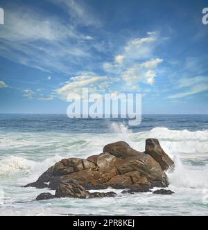 Copia lo spazio di un mare turbolento con maree modeste e onde instabili da forti venti che si infrangono su grandi massi della spiaggia con uno sfondo nuvoloso di cielo blu. Costa rocciosa a Western Cape, Sud Africa Foto Stock