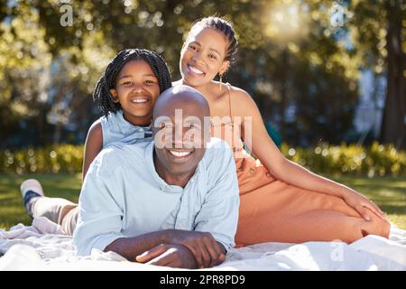 Ritratto felice famiglia afro-americana di tre persone che trascorrono del tempo di qualità insieme nel parco durante l'estate. Madre, padre e figlia che si legano insieme fuori. Una ragazza carina e genitori che sorridono all'aperto Foto Stock
