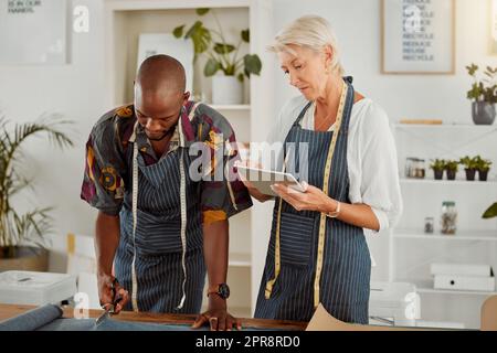 Due designer che utilizzano un tablet digitale mentre lavorano con il materiale al lavoro. Sarto caucasico maturo che parla e tiene in mano un tablet digitale mentre lavora con un collega afroamericano maschio. Uomo d'affari che taglia materiale Foto Stock