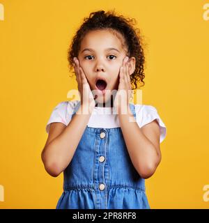 Ritratti in studio di una ragazza di razza mista che sembra scioccata con le mani sulle guance isolate su uno sfondo giallo. Carino bambino ispanico che si posa all'interno. Sorpreso e stupito ragazzo senza parole Foto Stock