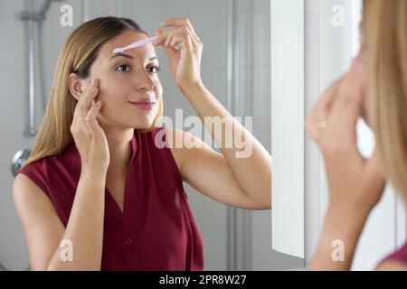 Donna ispanica che rasa le sopracciglia con il rasoio nello specchio di casa Foto Stock