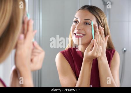 Bella giovane donna che rasa le sopracciglia con un rasoio sullo specchio a casa Foto Stock