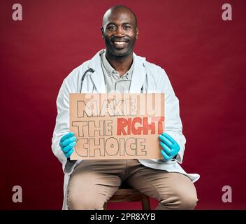 Medico afro-americano contro la covid che tiene e mostra un poster. Ritratto di un medico nero sorridente isolato su sfondo rosso dello studio con copyspace. Uomo che promuove e incoraggia il vaccino della corona in segno Foto Stock