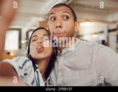 Primo piano di una giovane donna simpatica che tiene in mano il telefono cellulare mentre scatta un selfie con il suo ragazzo e fa facce in un bar. Amate coppie di razza mista che giocano mentre si siedono insieme a un appuntamento Foto Stock