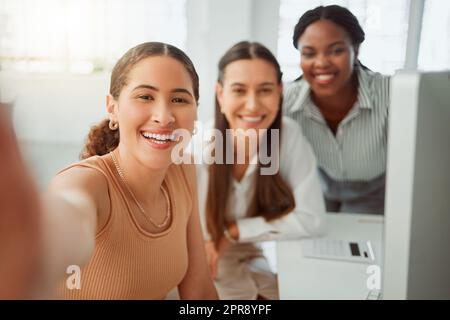 Ritratto di una giovane donna d'affari ispanica che scatta selfie con i suoi colleghi in un ufficio. Gruppo di tre donne sorridenti che scattano foto come un team ambizioso e dedicato in un'agenzia di startup creativa Foto Stock