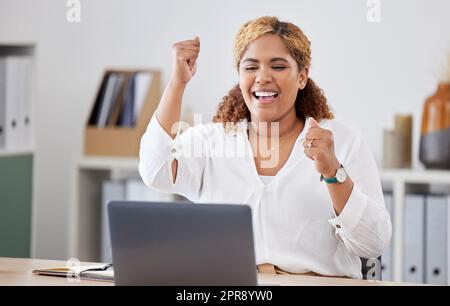 Una donna d'affari con gare miste che tira in allegria mentre usa un notebook da sola al lavoro. Una donna d'affari ispanica che sembra entusiasta mentre lavora su un computer a una scrivania in un ufficio Foto Stock