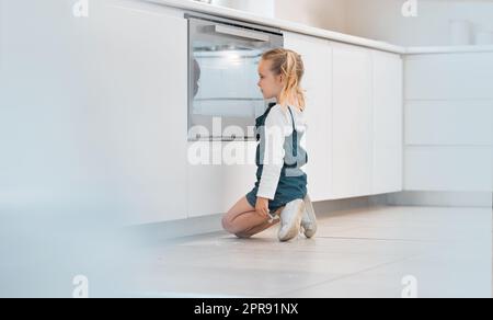 Bambina che aspetta pazientemente davanti al suo forno. Bambino caucasico che aspetta il suo cibo al forno. Bambina bionda che guarda il suo cibo che cuoce nel forno. Bambino che guarda nel forno Foto Stock