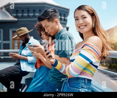 Ritratto di una giovane donna asiatica sorridente che si trova all'aperto con gli amici e che usa il suo cellulare per navigare in Internet. Gruppo di millennial diversi che utilizzano i loro telefoni per connettersi ai social media Foto Stock