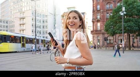 Donna ridente che tiene smartphone camminare su sfondo urbano. Guardando di lato. Visualizzazione ritagliata dell'intestazione. Foto Stock