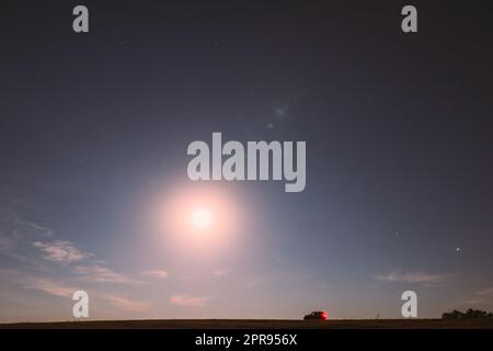 Luna piena che sorge sopra il campo in campagna. Vista notturna delle stelle naturali incandescenti. Auto parcheggiata su campo sotto Blue Night Starry Sky con Luna Foto Stock