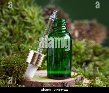 Flacone contagocce verde aperto su un pezzo di legno tra muschio verde, primo piano, mockup cosmetico naturale Foto Stock