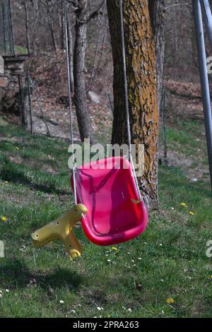 Oscillazione rotta su un parco giochi abbandonato con posto a sedere viola-giallo. Il sedile è rotto. L'erba e un albero sono visibili sullo sfondo. Foto Stock