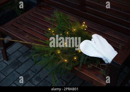 Rami di conifere decorati con una ghirlanda giacciono su una panchina insieme a guanti bianchi a maglia. Foto Stock