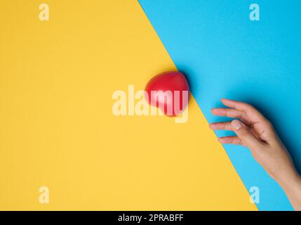 Il cuore rosso e la mano di una donna lo raggiunge su uno sfondo blu-giallo Foto Stock