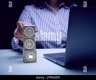 Una donna siede a un tavolo con un notebook e tiene in mano cubi di legno con icone. Foto Stock