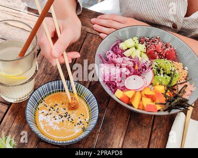 Donna che mangia gustoso colorato sano naturale vegetariano Hawaiiano poke ciotola utilizzando chopsticks asiatici su rustico tavolo di legno. Concetto sano di alimentazione naturale organica Foto Stock
